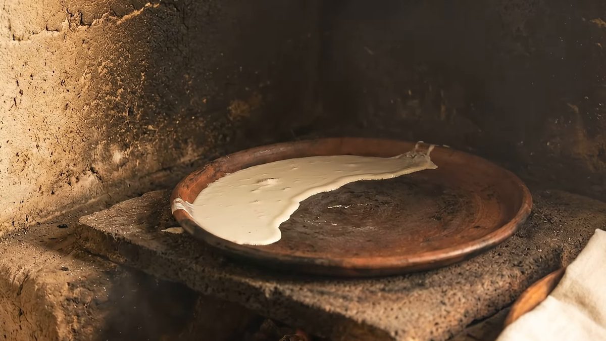Comal de barro sobre un fogón, con la masa extendida y pegada a la superficie.