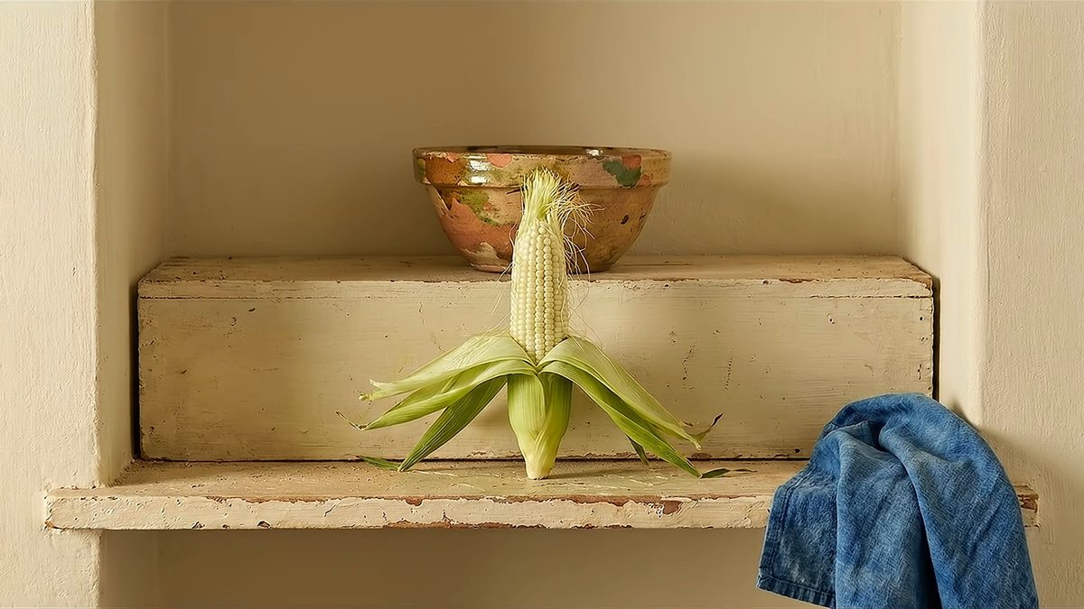 Elote con las hojas abiertas, de pie en un estante de pared encalada, junto a un cuenco de barro y un paño azul.