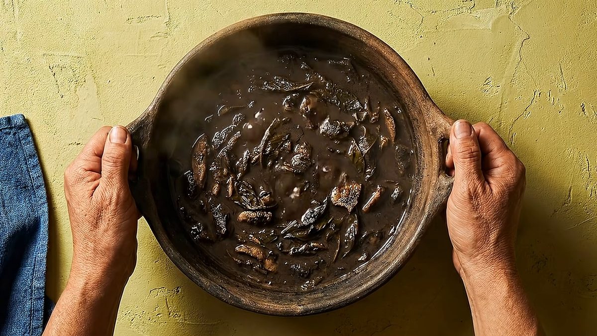 Dos manos sostienen una cazuela de barro con huitlacoche guisado en su caldo oscuro.