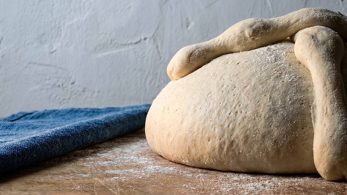Pan de muerto crudo, ya formado con sus tiras, sobre una mesa de madera junto a un paño azul.