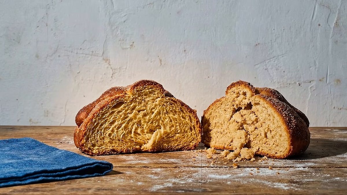 Pan de muerto partido en dos sobre una mesa de madera enharinada, junto a un paño azul.