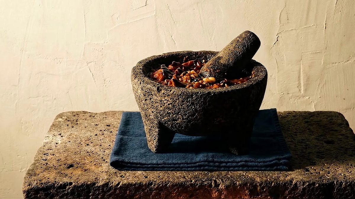 Molcajete de piedra volcánica lleno de salsa martajada, con el tejolote apoyado dentro, sobre un paño azul y una losa de piedra.