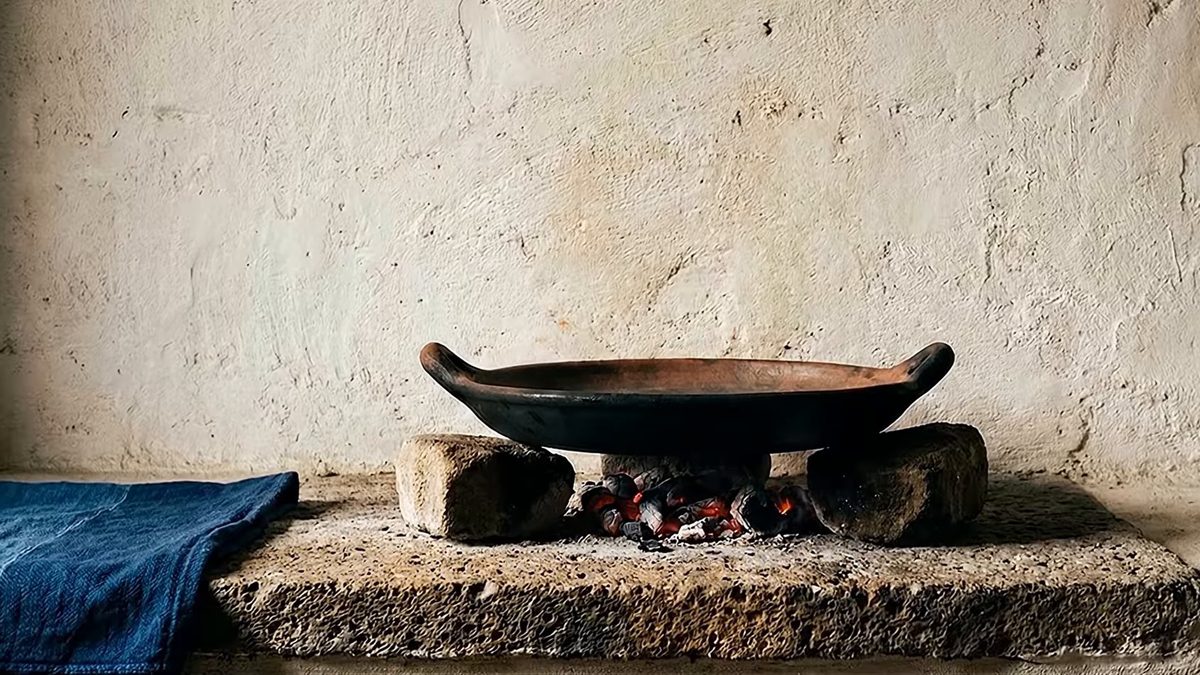 Comal de barro vacío sobre unas brasas, entre dos piedras, junto a un paño azul doblado.